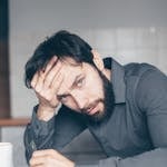Thoughtful man sitting at a table with glasses and cup, showing frustration.
